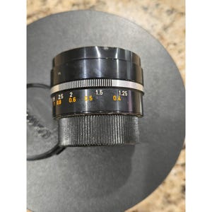 May include: A black camera lens with a silver ring and yellow and white markings. The lens has a ridged focus ring and is sitting on a dark gray surface. The lens has measurements in feet.