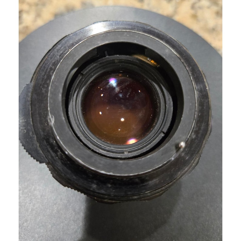 以下が含まれることがあります： 暗い金属製のハウジングを備えたカメラレンズのクローズアップ。レンズは反射性のマルチカラーの表面を持っています。ハウジングにはグリップ用のテクスチャリングと小さなレバーが付いています。全体的なデザインは、ヴィンテージまたはプロ仕様のカメラコンポーネントを示唆しています。