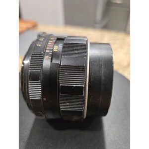 以下が含まれることがあります： リブと滑らかなセクションを備えた黒いカメラレンズ。レンズには白と赤のマーキングがあります。レンズは写真撮影と画像のキャプチャ用に設計されています。