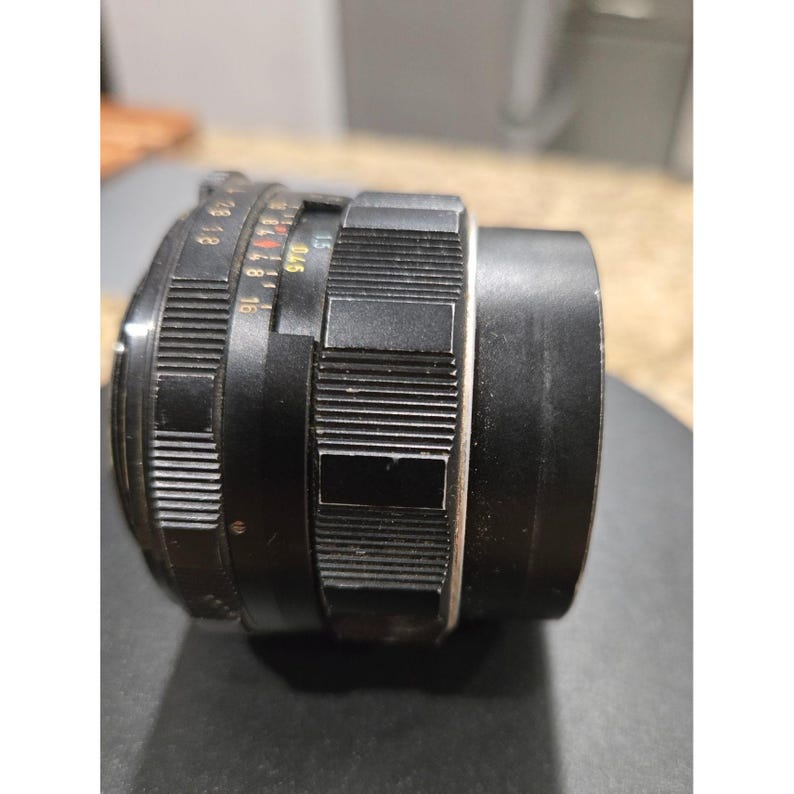 以下が含まれることがあります： リブと滑らかなセクションを備えた黒いカメラレンズ。レンズには白と赤のマーキングがあります。レンズは写真撮影と画像のキャプチャ用に設計されています。