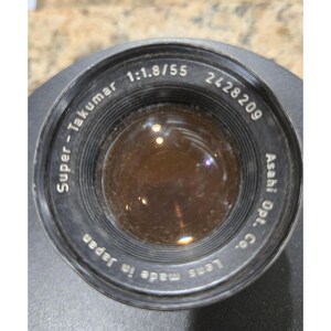 以下が含まれることがあります： 黒色のSuper-Takumar 1:1.8/55カメラレンズ。テキストには「Asahi Opt. Co.」と「Lens made in Japan」と記載されています。レンズのシリアル番号は2428209です。