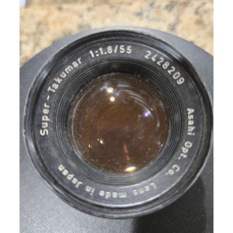 以下が含まれることがあります： 黒色のSuper-Takumar 1:1.8/55カメラレンズ。テキストには「Asahi Opt. Co.」と「Lens made in Japan」と記載されています。レンズのシリアル番号は2428209です。