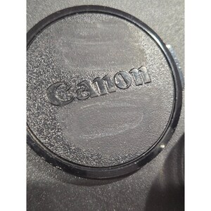 May include: A black Canon lens cap with the brand name embossed in the center. The cap has a textured surface and a circular design.