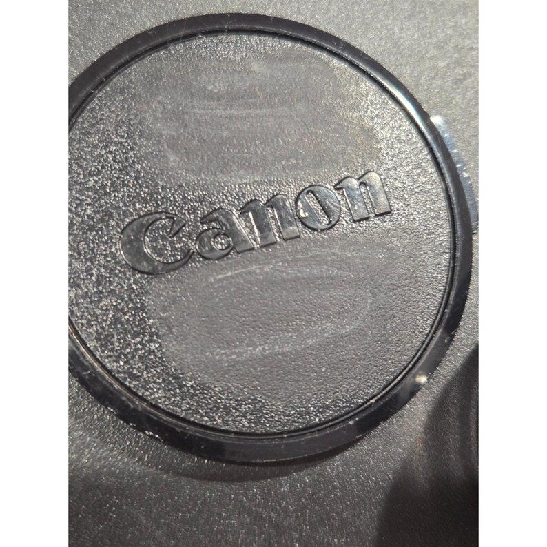 May include: A black Canon lens cap with the brand name embossed in the center. The cap has a textured surface and a circular design.