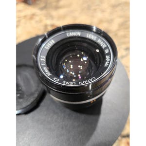 May include: A Canon EX 35mm f/3.5 camera lens. The lens is black with silver lettering and markings. The text "CANON LENS MADE IN JAPAN" is visible. The lens has a black lens cap.
