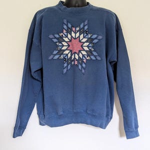 Op de afbeelding: Een blauw sweatshirt met lange mouwen en een grote, centraal geplaatste stervormige applicatie. De ster is opgebouwd uit diverse stoffen in blauwe, witte en roze tinten, wat een geometrisch, quiltachtig patroon op de voorzijde vormt.
