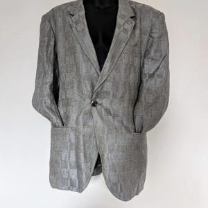 May include: A gray blazer with a checkered pattern, displayed on a black mannequin. The blazer has a notched lapel, two front pockets, and a single-button closure. The fabric appears to have a subtle texture.