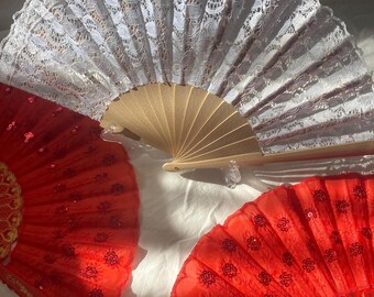 May include: Three decorative hand fans. One is white lace, and the other two are red with sequin and floral details. The fans have wooden handles and are spread open, showcasing their intricate designs. The fans are on a white surface.