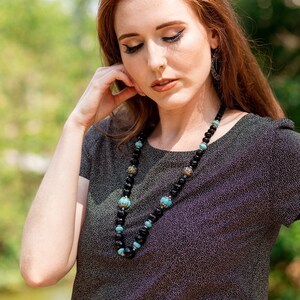 May include: A woman wearing a black sparkly top with a long necklace made of black beads and turquoise accents.