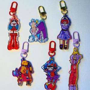 May include: A selection of acrylic keyrings with cartoon character designs. The keyrings feature a variety of characters, including a skeleton, a chess piece, a clown, and a purple monster, each with a different outfit and expression, and gold-coloured clasps.