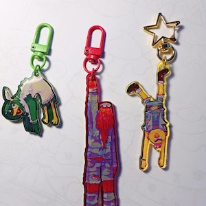 Może przedstawiać: Trzy kolorowe breloczki z postaciami z kreskówek. Pierwszy brelok jest zielony i przedstawia ptasie stworzenie z zielono-białym wzorem. Drugi brelok jest czerwony i przedstawia wysoką, rudowłosą postać z fioletowo-niebieskim wzorem. Trzeci brelok jest złoty i przedstawia żółtą postać z niebiesko-białym wzorem, stojącą do góry nogami.