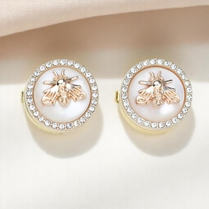May include: Two gold-toned, round earrings. Each earring features a white center with a gold bee design and a surrounding halo of small, clear crystals. The earrings are set against a soft, neutral background.