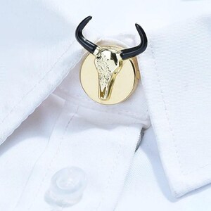 May include: A gold-colored bull skull cufflink with black horns is attached to a white dress shirt. The cufflink is round and features a detailed skull design. The shirt's button is visible in the lower part of the image.