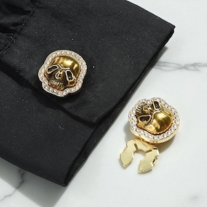 May include: Gold-colored skull cufflinks with a crystal border, set against a black shirt cuff. The cufflinks have a detailed skull design and a shiny finish, adding a touch of gothic flair.