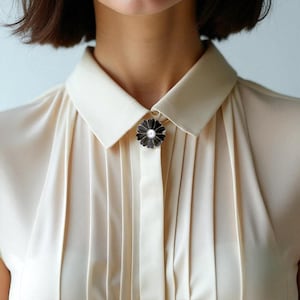 May include: A black and silver flower brooch with a pearl centre is pinned to a cream-coloured blouse. The brooch is a daisy shape, and the blouse has vertical pleats.