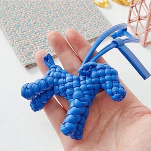 Puede incluir: Un llavero con forma de perro de cuero azul tejido. El llavero se sostiene en una mano, mostrando su intrincado diseño trenzado. El perro tiene un lazo para sujetar llaves y una correa.