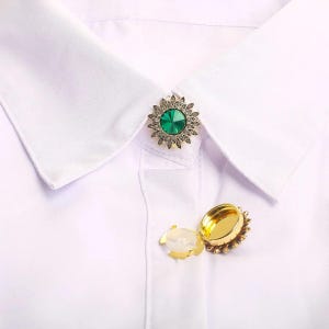 May include: A gold-coloured collar pin featuring a central emerald-green gemstone, encircled by a floral pattern. The pin is fastened to a white collared shirt. A matching open locket is also visible.