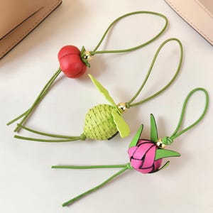 Puede incluir: Tres colgantes decorativos con cordones verdes y detalles dorados. Uno es un tomate rojo, otro una fruta verde tejida y el tercero una flor rosa. Los colgantes son probablemente para un bolso o llavero.