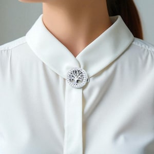 May include: A silver-coloured collar clip featuring a tree of life design, embellished with small, clear crystals. The clip is fastened to a white collared shirt, providing a decorative accent. The tree of life symbolises growth and connection.