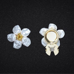 Peut inclure: Deux accessoires en forme de fleur à l'aspect perlé. L'un est une fleur complète avec un centre doré, et l'autre a une base dorée avec une zone circulaire. Les pétales sont blancs et de forme irrégulière.