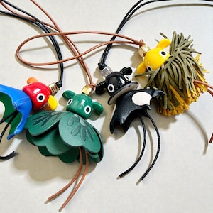 May include: A selection of quirky pendant necklaces. Each pendant showcases a distinctive creature design in blue, red, green, black, and yellow. The pendants are suspended from black and brown cords.