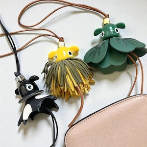 May include: Three whimsical bag charms. One is a black and white cow-like creature, another is a yellow creature with a green and yellow fringe, and the third is a green flower-like creature. All have brown leather cords.
