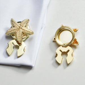 以下が含まれることがあります： 金色のヒトデ型カフスボタンセット。カフスボタンは、円形のベースの上に詳細なヒトデのデザインが施され、ユニークな留め金が付いています。白い背景にセットが表示され、カフスボタンのデザインと色が際立っています。