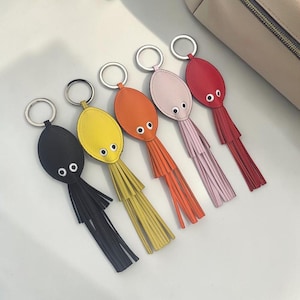 May include: A collection of five leather keychains in various colors: black, yellow, orange, pink, and red. Each keychain features a rounded oval shape with two small white eyes and a fringe of matching color. Silver key rings are attached.