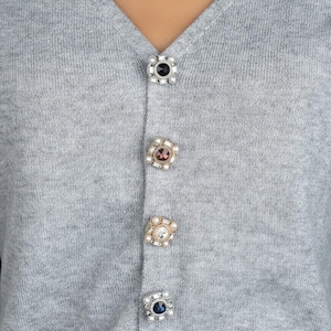 Puede incluir: Primer plano de un cárdigan gris con botones decorativos enjoyados. Los botones son cuadrados con piedras centrales de varios colores y detalles perlados. El cárdigan tiene cuello en V.