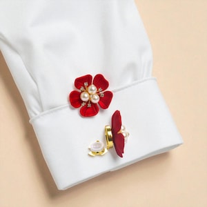 Può includere: Gemelli rossi a forma di fiore con perle e strass. Sono attaccati a un polsino di camicia bianca. I petali sono di colore rosso intenso, le perle bianche.