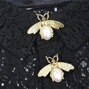 Puede incluir: Dos broches dorados con forma de abeja y centros en forma de perla se muestran sobre un fondo de encaje negro. Los broches tienen alas y antenas detalladas, creando un diseño delicado y elegante. El contraste entre el oro y el encaje negro añade interés visual.