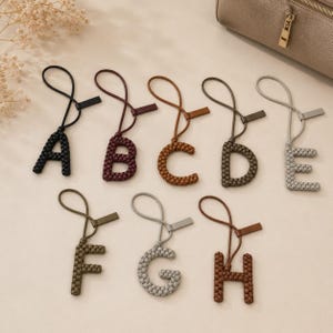 May include: A collection of eight letter-shaped bag charms, each crafted from woven material in various colours including black, burgundy, brown, olive green, grey, and tan. Each charm features a matching loop and a small rectangular tab attachment.