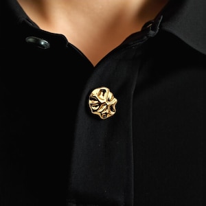 Peut inclure: Gros plan sur un bouton doré au design abstrait et texturé. Le bouton est fixé sur une chemise noire, créant un contraste entre le bouton brillant et le tissu sombre. La forme unique du bouton ajoute une touche d'élégance.
