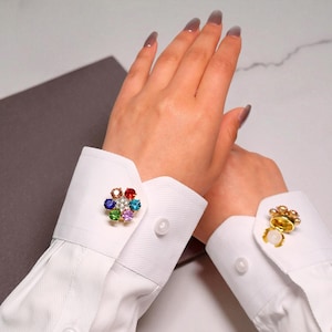 May include: Close-up of two white shirt cuffs with decorative cufflinks. One cufflink is a gold-toned flower shape with colourful gemstones. The other cufflink is a gold-toned flower shape with a white pearl.