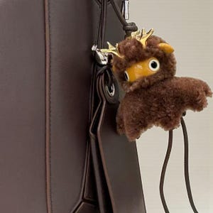 May include: A brown plush reindeer charm with gold antlers and yellow accents hangs from a dark brown leather handbag. The charm has large, round eyes and a fluffy texture, adding a whimsical touch to the bag.