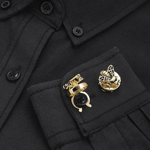 Puede incluir: Primer plano de una camisa negra con gemelos dorados. Un gemelo presenta un diseño de abeja y el otro tiene un diseño en forma de anillo. La camisa tiene botones negros.