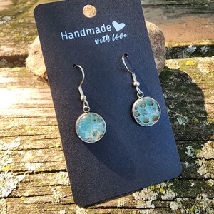May include: A pair of silver dangle earrings with round green and blue resin cabochons. The earrings are on a black card with the text "Handmade with love".