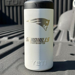 Puede incluir: Vaso aislado blanco con tapa negra. El vaso presenta el logotipo de Sandhills Patriots en dorado y la marca YETI. El dise&ntilde;o incluye un logotipo de Patriot estilizado.