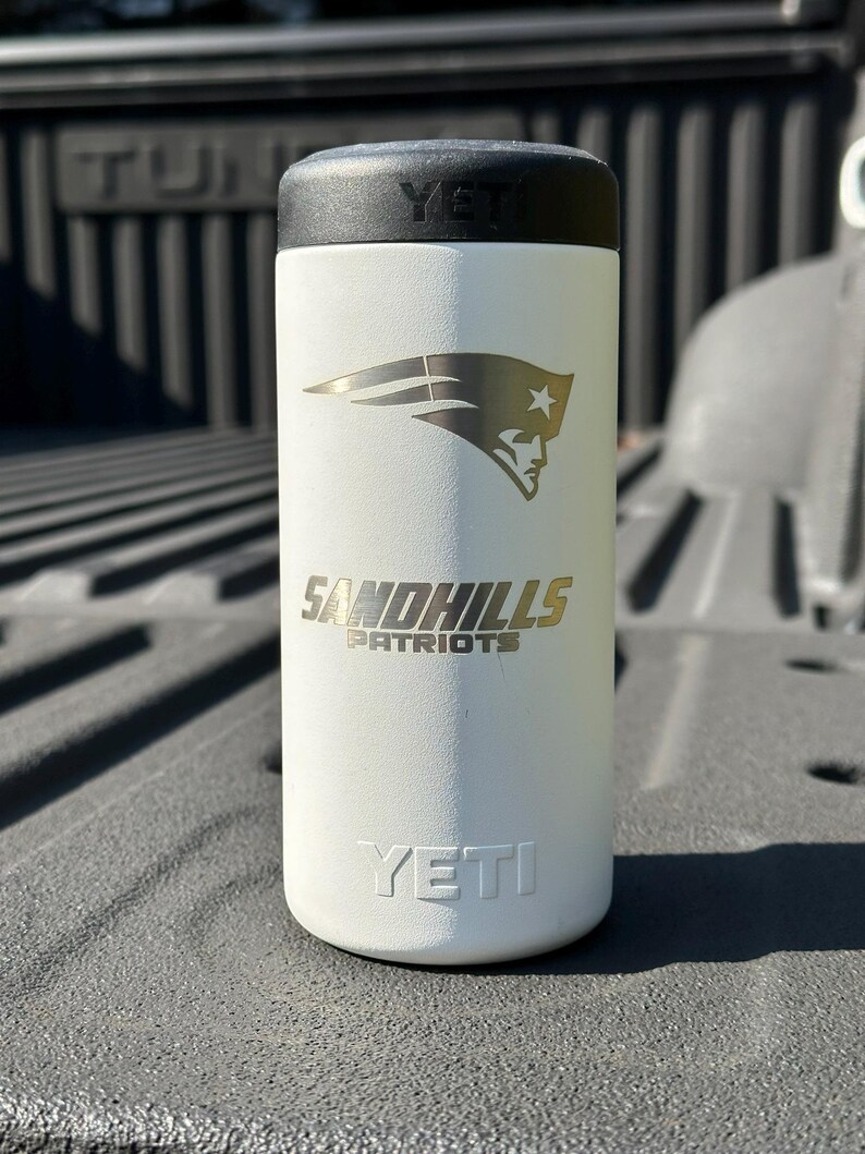 Puede incluir: Vaso aislado blanco con tapa negra. El vaso presenta el logotipo de Sandhills Patriots en dorado y la marca YETI. El dise&ntilde;o incluye un logotipo de Patriot estilizado.