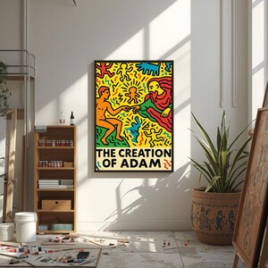 以下が含まれることがあります： キース・ヘリングの「アダムの創造」の額装されたアートプリント。このアートワークは、オレンジ、赤、青、黄色の鮮やかな人物が黒い背景に描かれています。タイトルは太字の白い文字で表示されています。プリントは明るいスタジオに展示されています。