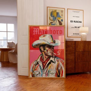 May include: A framed rectangular art print featuring a stylized painting of a person wearing a cowboy hat and smoking a cigarette. The background is split between red and pink, with the word Marlboro printed in white at the top of the image.