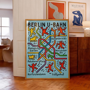 May include: A framed poster titled BERLIN U-BAHN featuring a light blue background with a colorful subway map design. The map includes intersecting lines in red, yellow, green, blue, and purple, interspersed with stylized dancing figures.