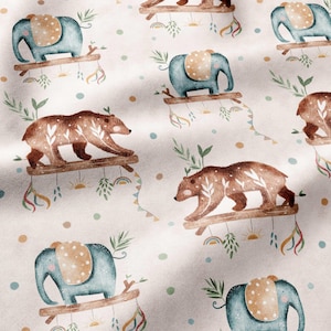 Op de afbeelding: Een stofpatroon met herhalende illustraties van blauwe olifanten en bruine beren, elk staand op een houten tak. Het ontwerp bevat kleine witte bladmotieven op de beren, stippen en hangende decoratieve elementen zoals regenbogen.