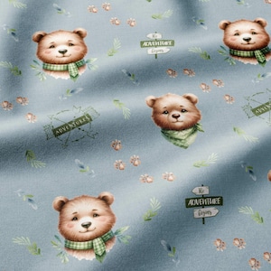 May include: A light blue fabric featuring a repeating pattern of brown bear heads wearing green plaid scarves, small paw prints, green leaves, and wooden signposts with the text "ADVENTURE BEGINS" printed in a dark green font.