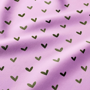 May include: A light purple fabric featuring a repeating pattern of small, dark green heart shapes. The material has visible folds and creases that create subtle shadows across the surface, highlighting the texture of the textile.