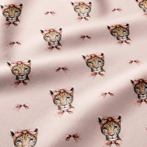 May include: A light pink fabric featuring a repeating pattern of leopard heads, each wearing a dusty rose headband and a matching bow at the neck, interspersed with small, simple dusty rose bows scattered across the soft-toned background.