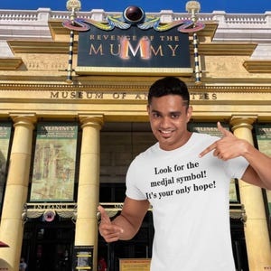 Puede incluir: Una camiseta blanca con texto negro que dice "¡Busca el símbolo medjal! ¡Es tu única esperanza!" La persona que lleva la camiseta está de pie frente a un edificio con las palabras "Revenge of the Mummy" y "Museum of Antiquities" en él.