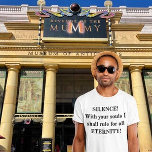 Puede incluir: Una persona con una camiseta blanca con el texto "SILENCE! With your souls I shall rule for all ETERNITY!" de pie frente a un edificio con el letrero "REVENGE OF THE MUMMY MUSEUM OF ANTIQUITIES".
