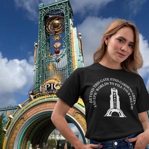 Puede incluir: Una camiseta negra con texto blanco que dice "Beyond this gate find gardens and epic worlds to fill your dreams". El texto está en un círculo alrededor de una silueta blanca de una torre del reloj. La camiseta la lleva puesta una persona de pie frente a una gran estructura metálica con la palabra "Epic" en ella.