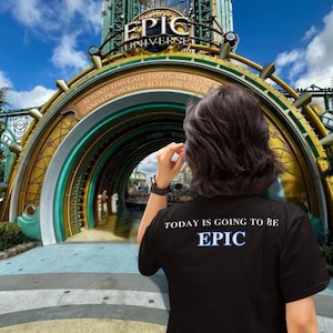 Puede incluir: Una persona lleva una camiseta negra con el texto "TODAY IS GOING TO BE EPIC" en letras blancas y un contorno azul. La persona está de pie frente a un arco grande y ornamentado con las palabras "EPIC UNIVERSE" en él.